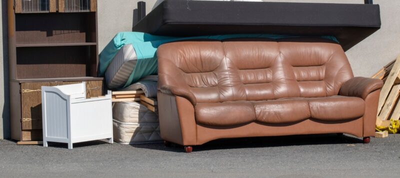 Old furniture and mattresses placed outside, including a brown leather sofa and a bookshelf