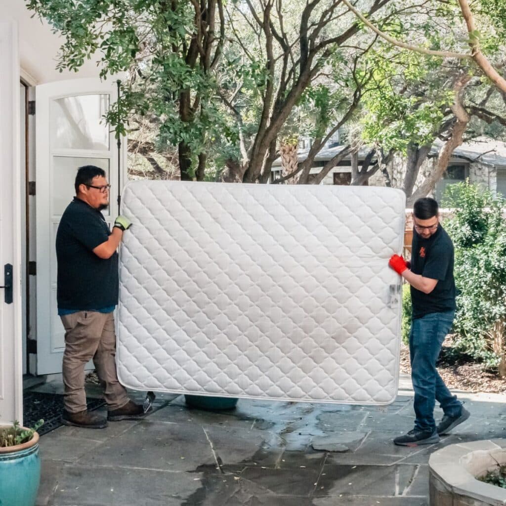 Two movers carrying a white mattress outside a house surrounded by trees