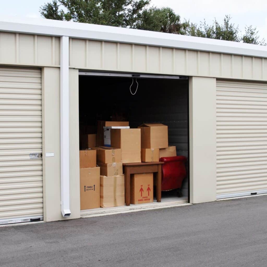 A storage unit with its door open, revealing stacked cardboard boxes and assorted furniture inside