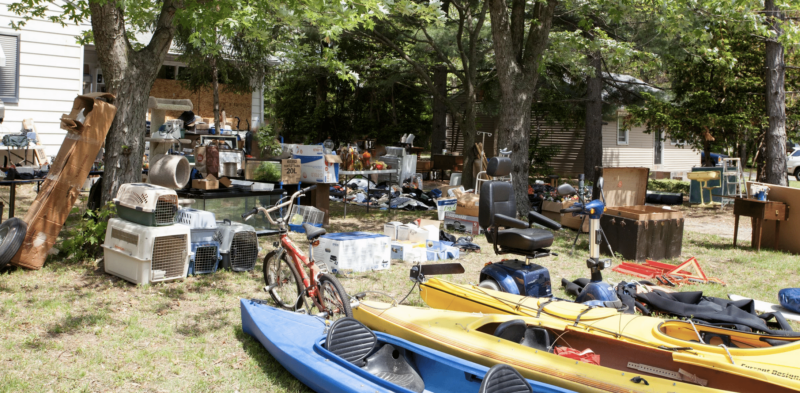Assorted items including kayaks, a bicycle, crates, and household goods spread across a cluttered yard during a property cleanout or estate sale